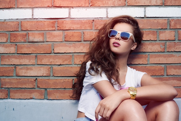 Schönes junges Mädchen in der Sonnenbrille auf einem Hintergrund einer Mauer. Schönes gesundes Haar. Jeans-Shorts