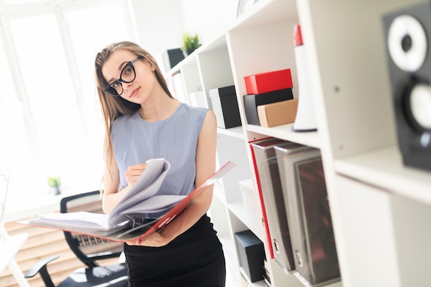 Schönes junges Mädchen in einem Büro steht nahe einem Gestell und rollt durch einen Ordner mit Dokumenten.