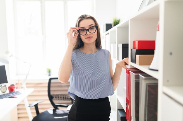 Schönes junges Mädchen in einem Büro steht nahe einem Regal mit Dokumenten und hält ihre Handgläser.