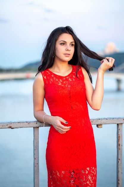 Schönes junges Mädchen in einem roten Kleid, das auf einer Brücke nahe einem alten Zaun aufwirft. Im Hintergrund sind ein Fluss, Brücken und Fragmente von Stadtgebäuden im Unschärfebereich zu sehen.