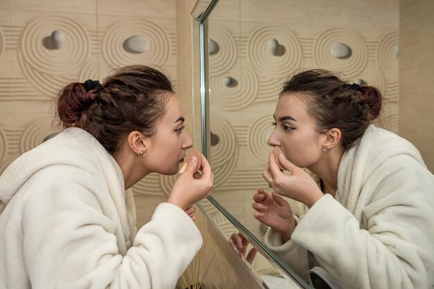 Schönes junges Mädchen in einem weißen Mantel macht ein langweiliges Make-up zu einem geräumigen Spiegel Das Konzept des Make-ups selbst