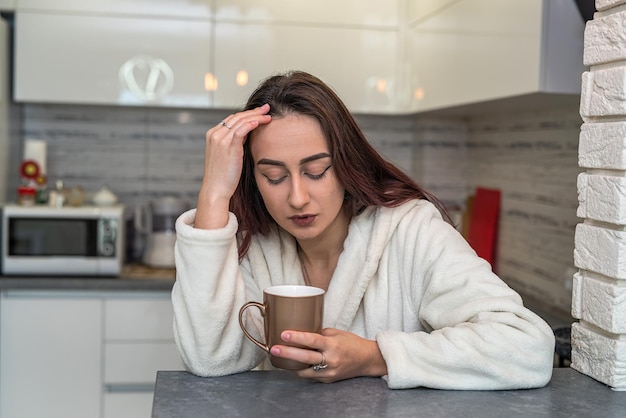 Schönes junges Mädchen in einem weißen Mantel und Turnschuhen, die morgens schläfrig sind, brüht Kaffee in ihrer eigenen Küche Schläfriges Frauenkonzept