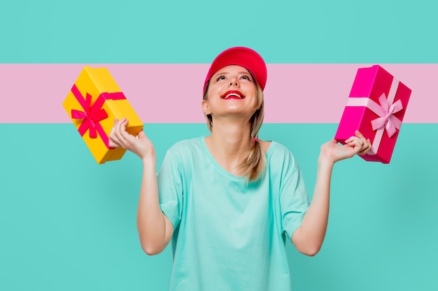 Schönes junges Mädchen in rosa Kappe und blauem T-Shirt mit Feiertagsgeschenkboxen auf grünem und rosa Hintergrund.