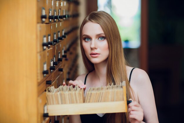 Schönes junges Mädchen mit dunklem glattem Haar öffnet Kasten und sucht nach Daten im Aktenschrank in der Bibliothek