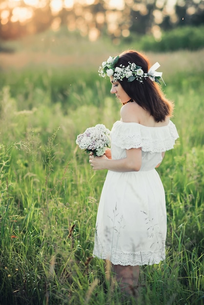 Schönes junges Mädchen mit einem Blumenstrauß und einem Kranz in einem weißen Kleid
