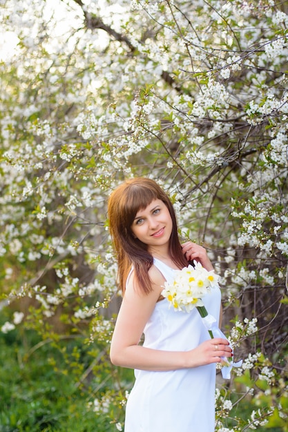 Schönes junges Mädchen mit einem Blumenstrauß von Narzissen in einem weißen Kleid auf einem Hintergrund von Kirschblüten