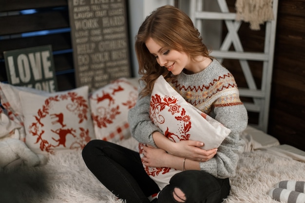 Schönes junges Mädchen mit einem Kissen in einem Vintage-Pullover sitzt auf dem Bett