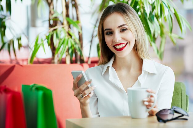 Schönes junges Mädchen mit hellbraunem Haar und roten Lippen, die weiße Bluse tragen und mit bunten Einkaufstüten und Tasse Kaffee sitzen, Handy halten.