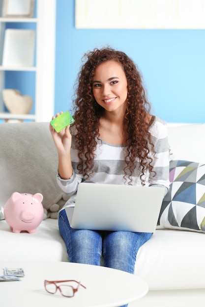 Schönes junges Mädchen mit Kreditkarte, Sparschwein und Laptop auf der Couch