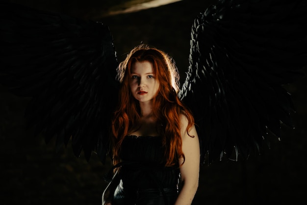Schönes junges Mädchen mit langen roten Haaren und schwarzen Flügeln in einem dunklen Raum