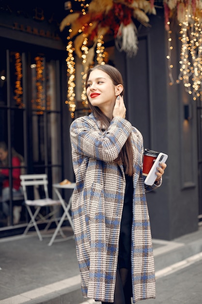 Schönes junges Mädchen mit roten Haaren, das draußen in einem Café steht und ein Telefon hält. Mädchen trinkt Kaffee. Mädchen hat rote Lippen und trägt einen stilvollen blauen Mantel.