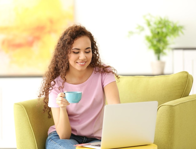 Schönes junges Mädchen mit Tasse Tee und Laptop auf Sessel