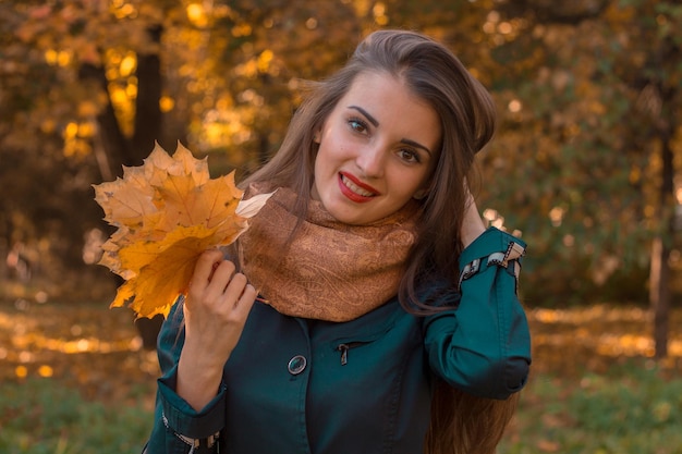 Schönes junges Mädchen steht und lächelt im Park und hält die Haare mit einer Hand, während die andere Hand die Blätter in der Nähe hält