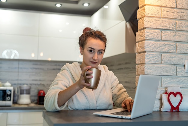 Schönes junges Mädchen wachte morgens auf, trank Kaffee in der Küche und arbeitete während einer Pandemie aus der Ferne Das Konzept der Arbeit während einer Pandemie Fernarbeit