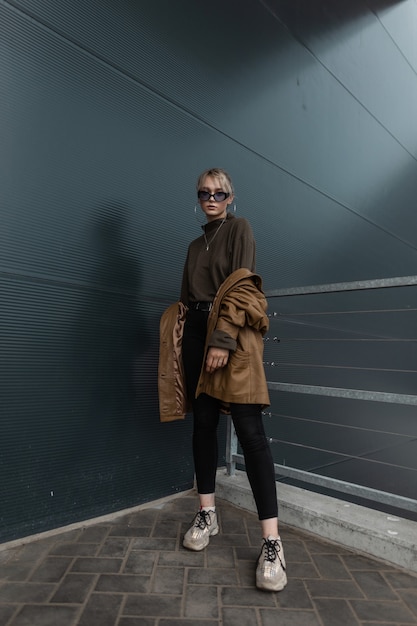 Schönes junges Mädchenmodell in einer modischen Lederjacke mit schwarzen Jeans und Turnschuhen in Sonnenbrille in der Nähe einer modernen Metallwand in der Stadt