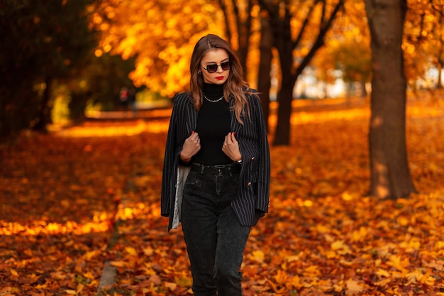 Schönes junges Model-Mädchen in einem eleganten Anzug mit einem schwarzen Pullover und einem Blazer geht im Park mit Herbstlaub