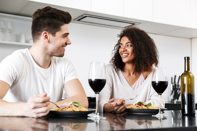 Schönes junges multiethnisches Paar, das ein romantisches Abendessen zu Hause hat, Rotwein trinkt und Pasta isst