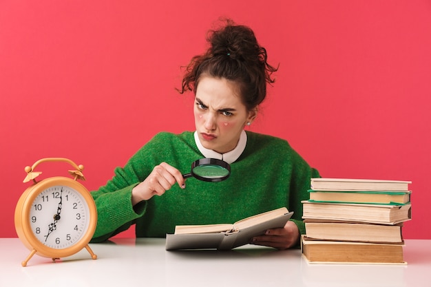 Schönes junges Nerd-Studentenmädchen, das am Tisch sitzt, isoliert, mit Büchern studierend, Lupe