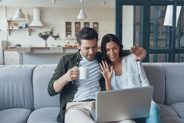 Schönes junges Paar, das Kaffee trinkt und auf dem Laptop schaut, während es zu Hause auf dem Sofa sitzt