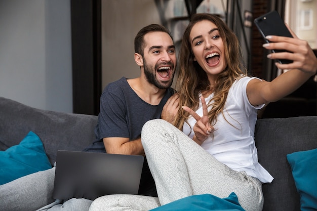 Schönes junges Paar, das sich auf einer Couch zu Hause entspannt, mit Laptop-Computer, ein Selfie nehmend