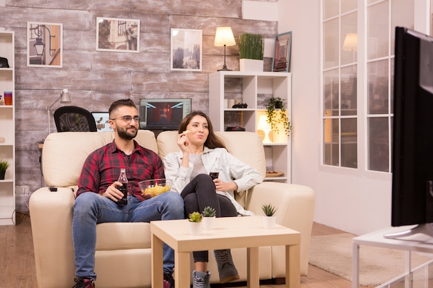 Schönes junges Paar, das Soda trinkt und Chips isst, während es einen Film im Fernsehen sieht, das auf der Couch sitzt.