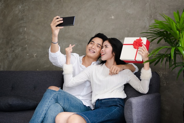 Schönes junges Paar hält Geschenkbox, tut selfie und lächelt beim zu Hause feiern