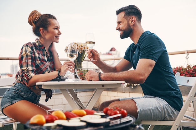 Schönes junges Paar in Freizeitkleidung, das ein romantisches Abendessen genießt und lächelt, während es draußen auf der Dachterrasse sitzt?