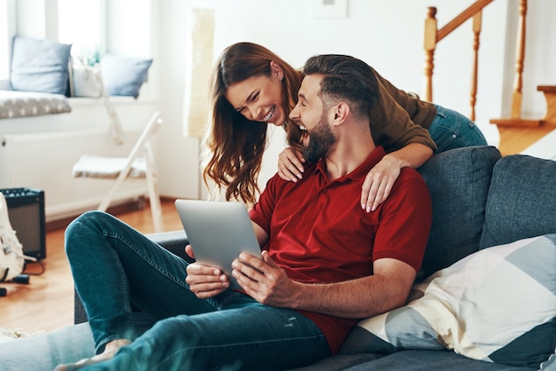 Schönes junges Paar in Freizeitkleidung, das mit jemandem spricht, der ein digitales Tablet verwendet, während er sich drinnen auf dem Sofa ausruht