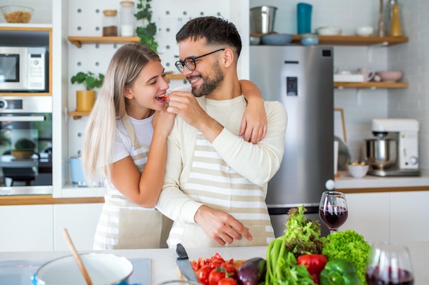 Schönes junges Paar sieht sich an und füttert sich gegenseitig mit einem Lächeln, während es zu Hause in der Küche kocht Liebevolles, fröhliches junges Paar, das sich umarmt und zusammen kocht und Spaß in der Küche hat