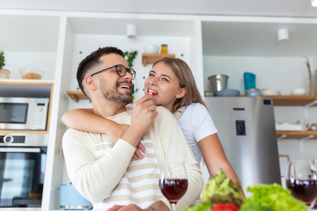 Schönes junges Paar sieht sich an und füttert sich gegenseitig mit einem Lächeln, während es zu Hause in der Küche kocht Liebevolles, fröhliches junges Paar, das sich umarmt und zusammen kocht und Spaß in der Küche hat