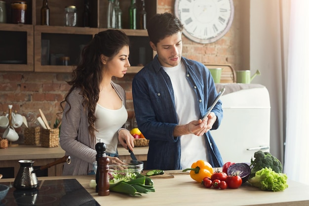 Schönes junges Paar verwendet digitale Tablette und liest Rezepte auf der kulinarischen Website, während es in der Loftküche zu Hause kocht, Platz kopieren