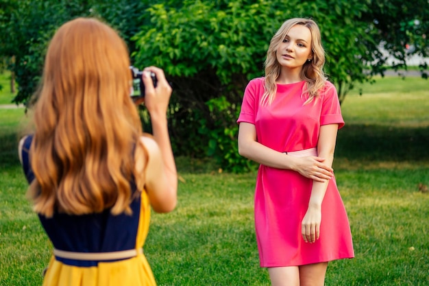 Schönes junges rothaariges irisches Mädchen in einem gelben Kleid und europäische blonde Frau in rosafarbenem Kleid fotografierten sich gegenseitig im Sommerpark. Fotoshooting von zwei Model-Freundinnen.
