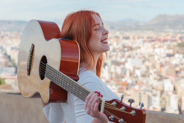 Schönes junges rothaariges Mädchen geht mit ihrer Gitarre im Freien, die Panoramablick auf die Stadt hat.