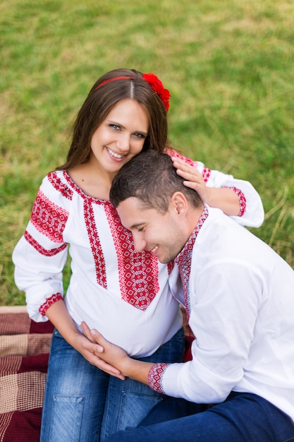 Schönes junges schwangeres Paar gekleidet im nationalen ukrainischen Stil, der Picknick im Herbstpark hat. Mutterschafts- und Familienglückskonzept.