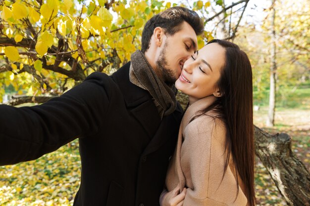 Schönes junges verliebtes Paar, das im Herbst Zeit zusammen im Park verbringt, ein Selfie macht, sich küssen