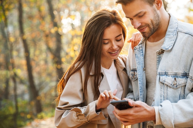 Schönes junges verliebtes Paar mit Handy, während es Zeit im Herbstpark verbringt