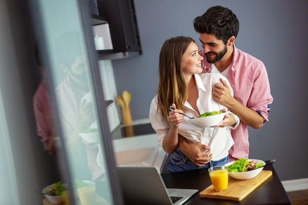 Schönes junges verspieltes Paar, das zusammen Salat in der Küche isst