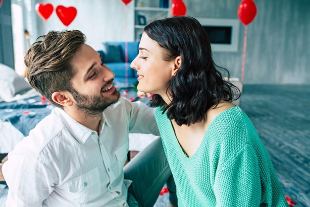 Schönes junges zartes Paar küsst und umarmt sich, während es auf dem Boden des Schlafzimmers mit romantischem Hintergrund sitzt. Valentinstag. Jubiläum. Datum. Liebeskonzept