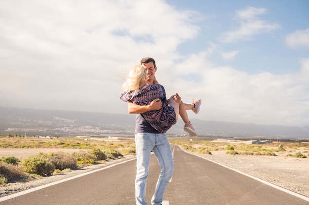 Schönes kaukasisches Paar genießt und hat Spaß am Sommertag. schöne Leute. der Mann trägt das blonde Mädchen. lange Asphaltstraße im Hintergrund und überall Wüste. blauer Himmel mit Wolken