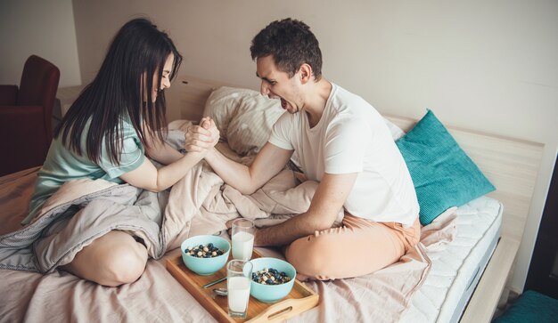 Schönes kaukasisches Paar spielt im Bett, bevor es Müsli mit Milch isst, die sich glücklich fühlt und ihre Kraft misst