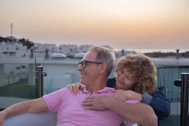 Schönes kaukasisches reifes Senior-Paar auf der Terrasse des Hauses, das den Sonnenuntergang über dem Meer genießt und sich kuschelt