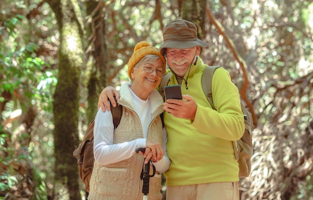 Schönes kaukasisches Seniorenpaar mit Rucksack, das zusammen im Wald wandert und einen gesunden Lebensstil und Ruhestand genießt, während es das Handy benutzt
