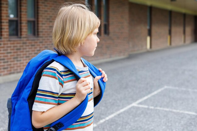 Foto schönes kind in der nähe des schulgebäudes auf dem schulhof, kleiner blonder schüler mit blauem rucksack