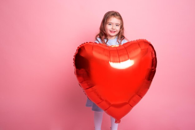 Schönes Kindermädchen mit rotem Herzballon auf rosafarbenem Hintergrund, der den Valentinstag feiert