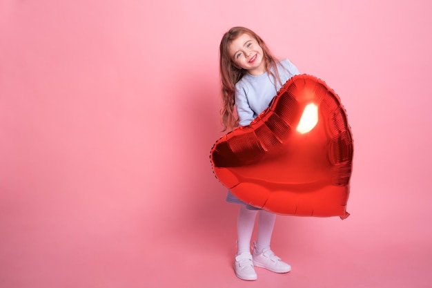 Schönes Kindermädchen mit rotem Herzballon auf rosafarbenem Hintergrund, der den Valentinstag feiert