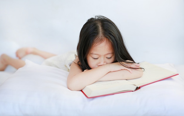 Schönes kleines asiatisches Kindermädchen, das das gebundene Buch liest mit Kissen auf weißem Hintergrund liest.