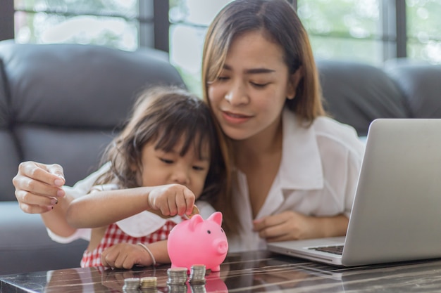 Schönes kleines asiatisches Mädchen und Mutter, die Geld in Sparschwein setzen, um zu sparen