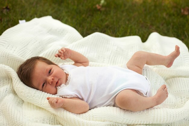 Schönes kleines Baby in weißer Kleidung, das auf der weißen Decke auf dem grünen Gras liegt