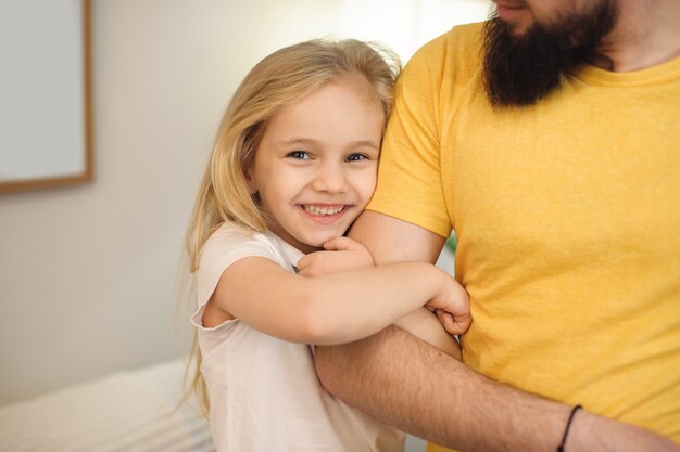 Schönes kleines blondes Mädchen, das lächelt, während es am Morgen die Hand ihres Vaters zu Hause umarmt.