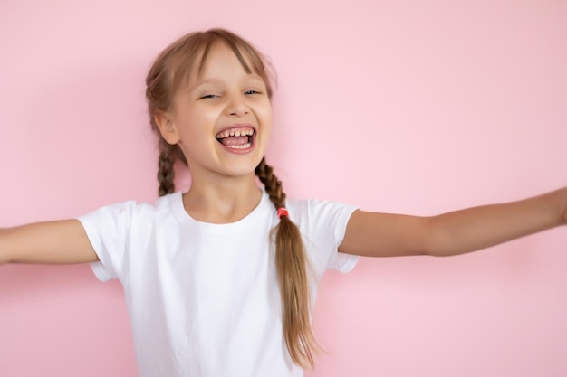 Schönes kleines blondes Mädchen mit langen Haaren im weißen T-Shirt, das auf einem rosa Hintergrund lächelt